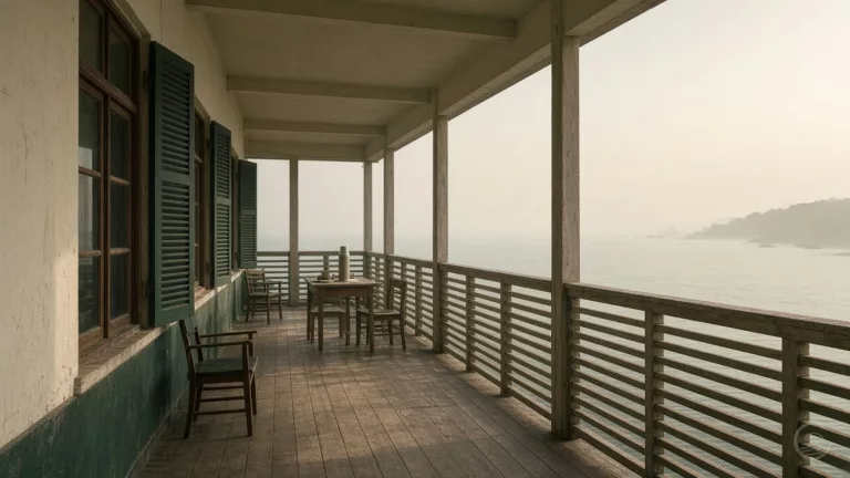 The long wooden veranda of an old seaside building with green shutters, wooden chairs and a small table with a thermos flask and teacups, looking out over a hazy, calm sea towards a wooded headland.